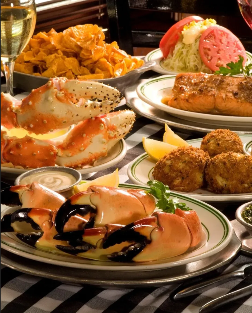 5 best restaurants in miami worth the trip | local picks 2026 4 A table set with plates of crab claws, crab cakes, salmon, shredded salad with tomato slices, a bowl of potato chips, lemon wedges, dipping sauces, and glasses of white wine on a checkered tablecloth.