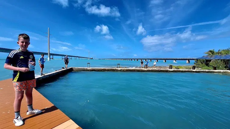 A boy stands on a wooden dock holding a drink near turquoise water, with a scenic ocean, blue sky, people in the background, and a long bridge stretching across the water.