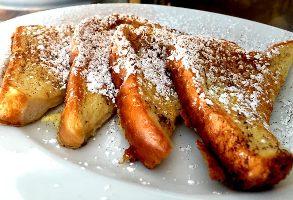 Four slices of golden-brown french toast on a white plate, dusted with powdered sugar and a sprinkle of cinnamon.