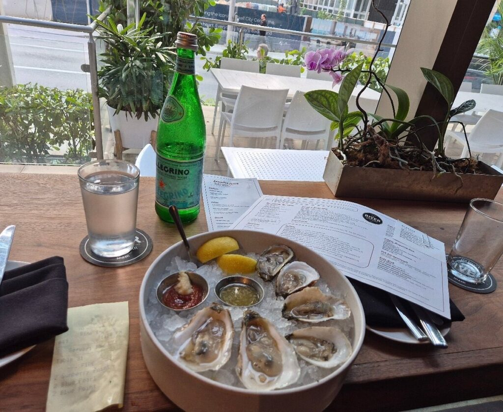 5 best restaurants in miami worth the trip | local picks 2026 7 A bowl of oysters with lemon wedges, sauces, and a bottle of san pellegrino sit on a wooden table by a window, alongside menus, a glass of water, utensils, and a notepad, with a street view outside.