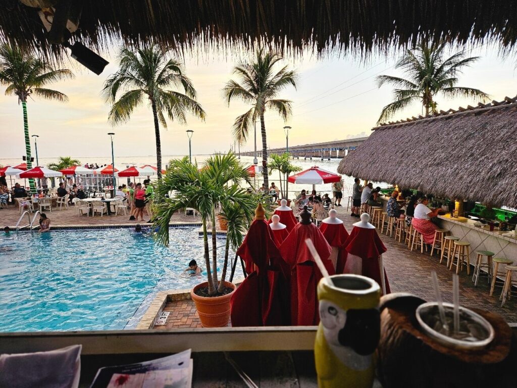 Florida keys happy hour: best overseas highway deals 4 View of a lively beachside bar and pool area with palm trees, red umbrellas, people swimming and sitting at tables, and drinks in the foreground under a thatched roof—capturing the vibrant atmosphere of a classic florida keys happy hour.