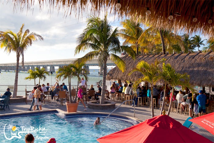 Florida keys happy hour: best overseas highway deals 5 People relax by a pool and tiki bar at a tropical beachside venue with palm trees, a bridge in the background, and the ocean beyond. Some are sitting, swimming, or standing under thatched roofs and red umbrellas, enjoying a classic florida keys happy hour as the sun sets over paradise.
