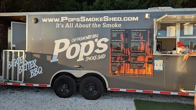 A gray food truck labeled pops smoke shed with bbq menu boards, a serving window, and the slogan its all about the smoke parked on gravel with trees in the background.