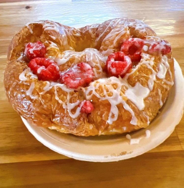 A heart-shaped pastry topped with icing and raspberries sits on a white plate, placed on a wooden table.