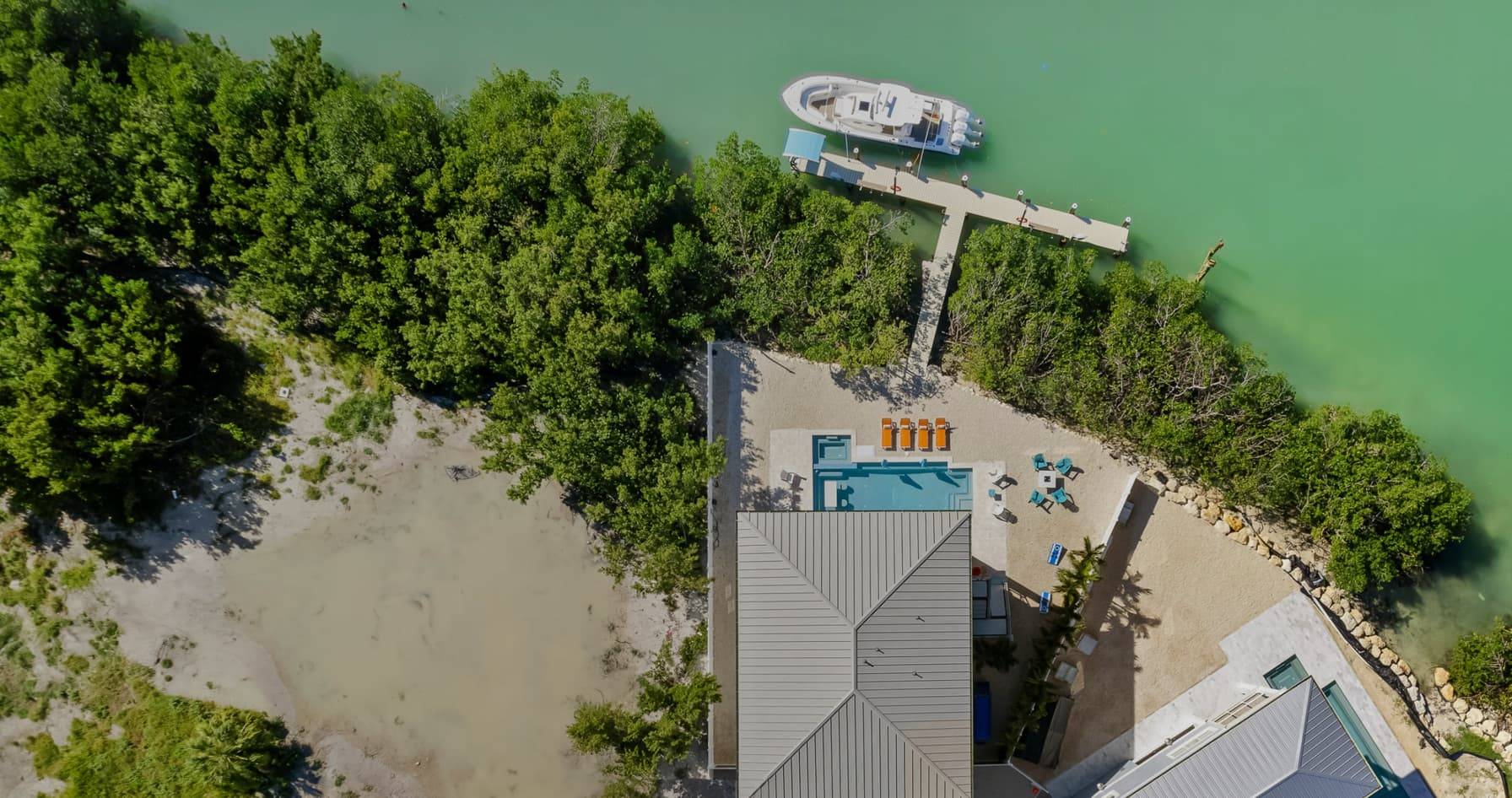 Aerial view of a waterfront property with a pool, lounging area, and docked boat, surrounded by lush greenery